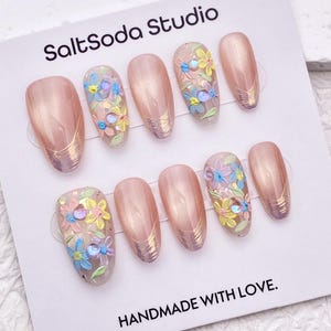 May include: A set of ten artificial nails in a light pink hue, some adorned with delicate floral designs in pastel colours. Gold accents and the text "SaltSoda Studio" and "HANDMADE WITH LOVE" are visible on the card.