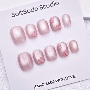 Könnte beinhalten: Ein Set aus perlmuttrosa Kunstnägeln auf einer weißen Karte. Einige Nägel haben kleine, erhabene rosa Verzierungen. Auf der Karte steht "SaltSoda Studio" und "HANDMADE WITH LOVE."