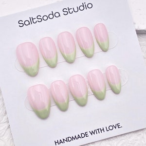 Pode incluir: Um conjunto de dez unhas postiças rosa e verde. As unhas são de forma oval e têm uma ponta verde claro. As unhas estão dispostas em duas filas em um cartão branco com o texto "SaltSoda Studio" e "HANDMADE WITH LOVE."
