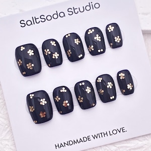 May include: A set of navy blue press-on nails with gold glitter flower accents. The nails are arranged on a white card with the text "SaltSoda Studio" and "HANDMADE WITH LOVE."