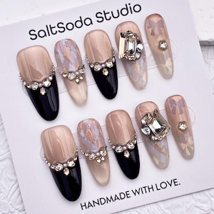 May include: A set of ten artificial nails with various designs. Some nails are black, nude, or adorned with rhinestones and bows. The card reads "SaltSoda Studio" and "HANDMADE WITH LOVE."