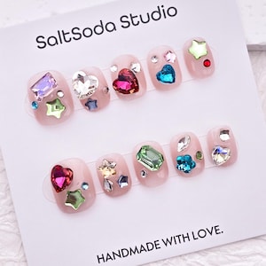 May include: A set of artificial nails with a light pink base, adorned with various colorful rhinestones in shapes like hearts, stars, and geometric forms. The card reads "SaltSoda Studio" and "HANDMADE WITH LOVE."