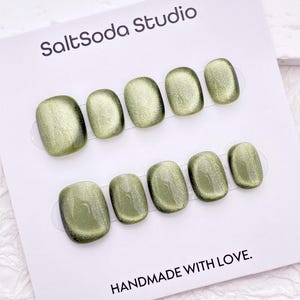 Könnte beinhalten: Ein Set olivgrüner Press-On-Nägel mit Cat-Eye-Effekt, angeordnet auf einer weißen Karte. Die Karte zeigt oben den Text "SaltSoda Studio" und unten "HANDMADE WITH LOVE". Die Nägel haben ein glänzendes Finish.