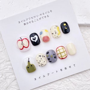May include: A set of ten false fingernails with different designs. The designs include a red apple, a black heart, a blue eye, a pink heart, a yellow and black polka dot, a black and white polka dot, a gray and green polka dot, a yellow and white plaid, a white nail, and a green pear.