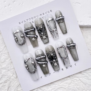 Puede incluir: Un conjunto de diez uñas postizas plateadas y grises con diferentes diseños de cadena plateada y metal. Las uñas se muestran en una tarjeta blanca con texto japonés que dice "Estilo de accesorios para uñas".