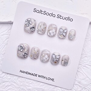 May include: A set of press-on nails with a light gray base, adorned with silver floral and butterfly designs, and gold star accents. The card reads "SaltSoda Studio" and "HANDMADE WITH LOVE."