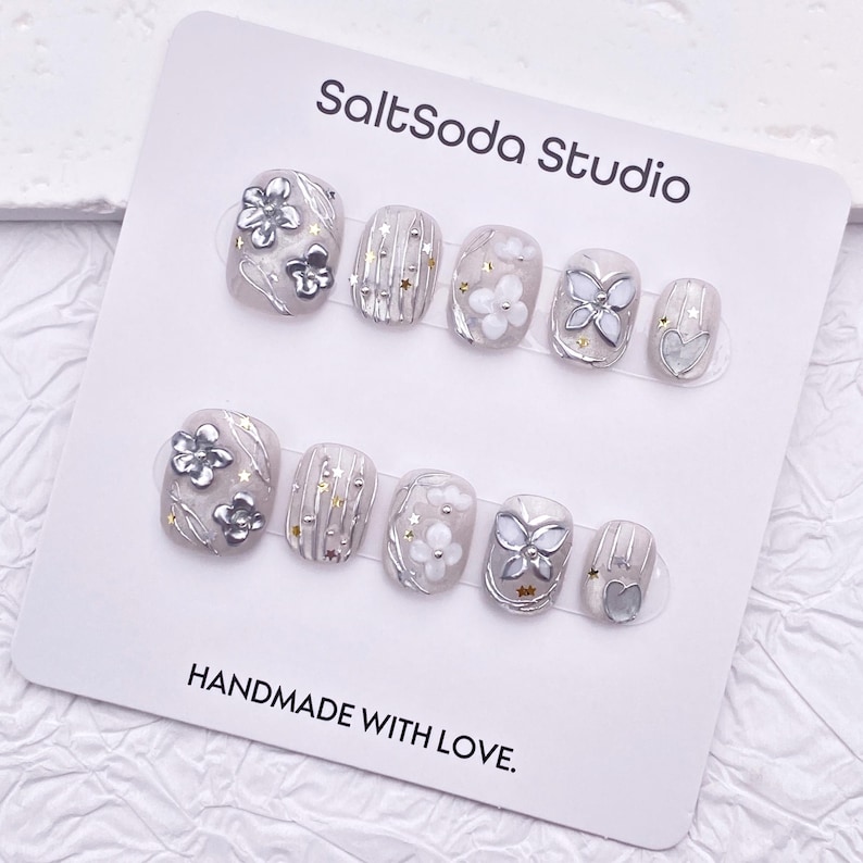 May include: A set of press-on nails with a light gray base, adorned with silver floral and butterfly designs, and gold star accents. The card reads "SaltSoda Studio" and "HANDMADE WITH LOVE."