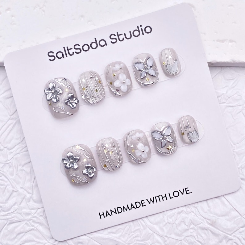 May include: A set of press-on nails from SaltSoda Studio. The nails are oval-shaped with a light gray base and feature silver floral and butterfly designs, along with gold star accents. The card reads "HANDMADE WITH LOVE."