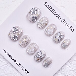 May include: A set of press-on nails in various shapes, featuring silver floral and star accents on a light gray base. The nails are decorated with white and silver embellishments. The text "SaltSoda Studio" and "HANDMADE WITH LOVE" are visible.