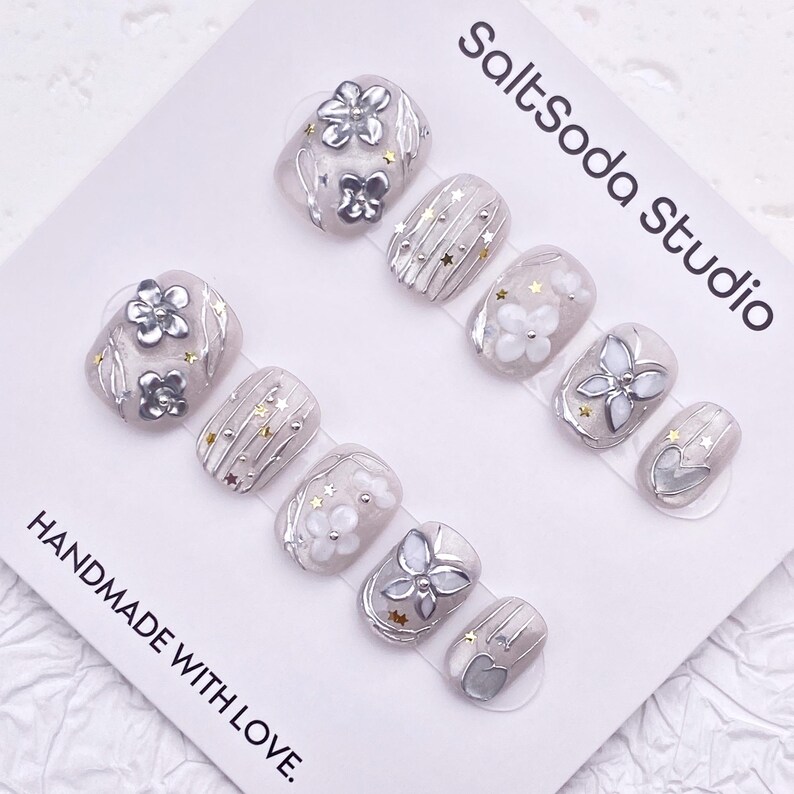 May include: A set of press-on nails in various shapes, featuring silver floral and star accents on a light gray base. The nails are decorated with white and silver embellishments. The text "SaltSoda Studio" and "HANDMADE WITH LOVE" are visible.