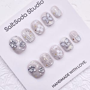 May include: A set of press-on nails with a light gray base, adorned with silver floral and star accents. The nails feature various designs, including embossed flowers and metallic embellishments. The text "SaltSoda Studio" and "HANDMADE WITH LOVE" are visible.