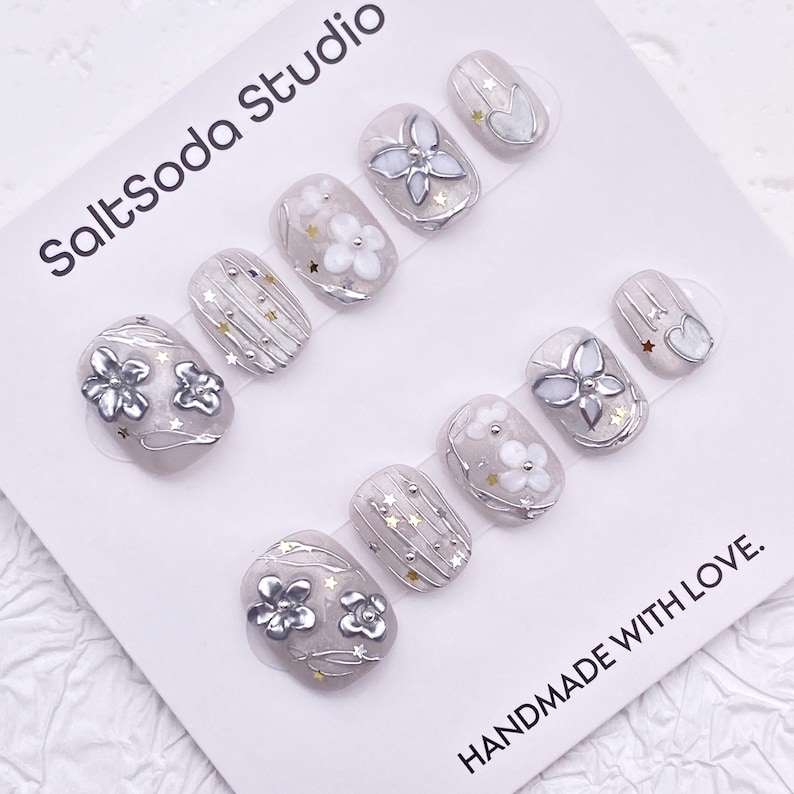 May include: A set of press-on nails with a light gray base, adorned with silver floral and star accents. The nails feature various designs, including embossed flowers and metallic embellishments. The text "SaltSoda Studio" and "HANDMADE WITH LOVE" are visible.