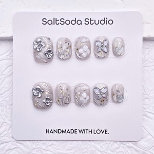 May include: A set of ten artificial nails in a variety of designs. The nails are primarily gray with silver and white floral and geometric accents. The text "SaltSoda Studio" and "HANDMADE WITH LOVE" are printed on the card.