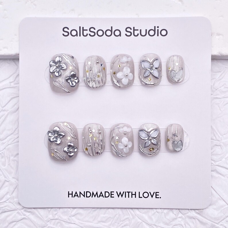 May include: A set of ten artificial nails in a variety of designs. The nails are primarily gray with silver and white floral and geometric accents. The text "SaltSoda Studio" and "HANDMADE WITH LOVE" are printed on the card.