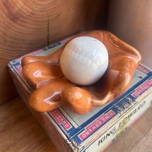 Puede incluir: Un plato vintage de cerámica con forma de guante de béisbol en un tono marrón anaranjado brillante sostiene una bola blanca con superficie agrietada. El plato descansa sobre una caja de cigarros King Edward Seventh vintage.