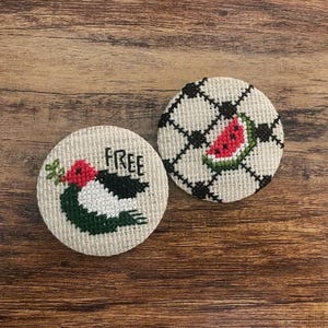 Könnte beinhalten: Zwei runde, bestickte Untersetzer. Einer zeigt einen schwarz-weißen Vogel mit dem Wort "FREE" und floralen Akzenten. Der andere hat eine Wassermelonen-Scheibe auf einem schwarz-weißen Karomuster. Beide liegen auf einer Holzoberfläche.
