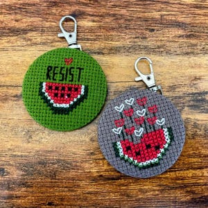 May include: Two circular cross-stitch keyrings. One is green with the word "RESIST" and a watermelon slice. The other is grey with a watermelon slice and red and white floral design. Both have silver clasps.