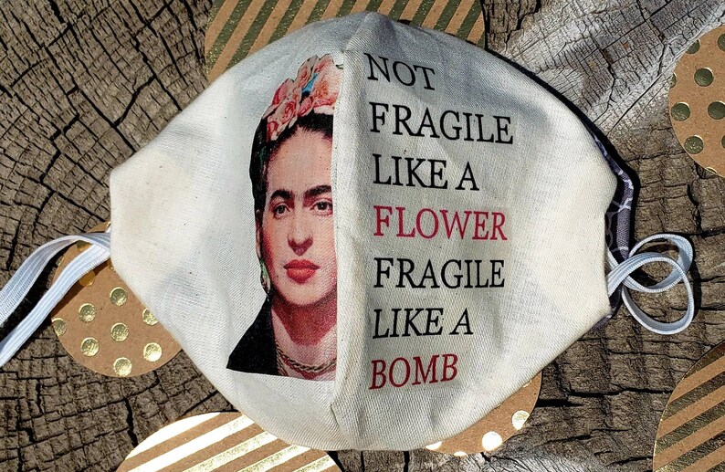 以下が含まれることがあります： 片側にフリーダ・カーロの絵、もう片側に「NOT FRAGILE LIKE A FLOWER FRAGILE LIKE A BOMB」というテキストが書かれた白いフェイスマスクです。