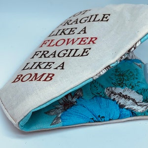 以下が含まれることがあります： 黒い文字と赤い文字で「Not Fragile Like A Flower Fragile Like A Bomb」と書かれた白いキャンバス地のポーチ。ポーチの内側は青い花柄の生地で裏打ちされている。