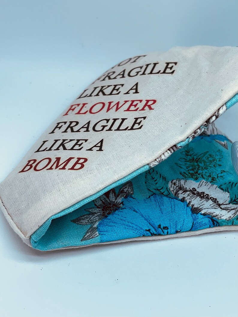 以下が含まれることがあります： 黒い文字と赤い文字で「Not Fragile Like A Flower Fragile Like A Bomb」と書かれた白いキャンバス地のポーチ。ポーチの内側は青い花柄の生地で裏打ちされている。