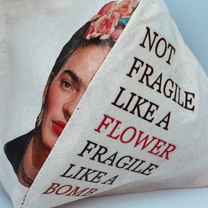 以下が含まれることがあります： フリダ・カーロのカラフルな画像と「Not Fragile Like a Flower Fragile Like a Bomb」というテキストが書かれた白いキャンバス地のトートバッグです。