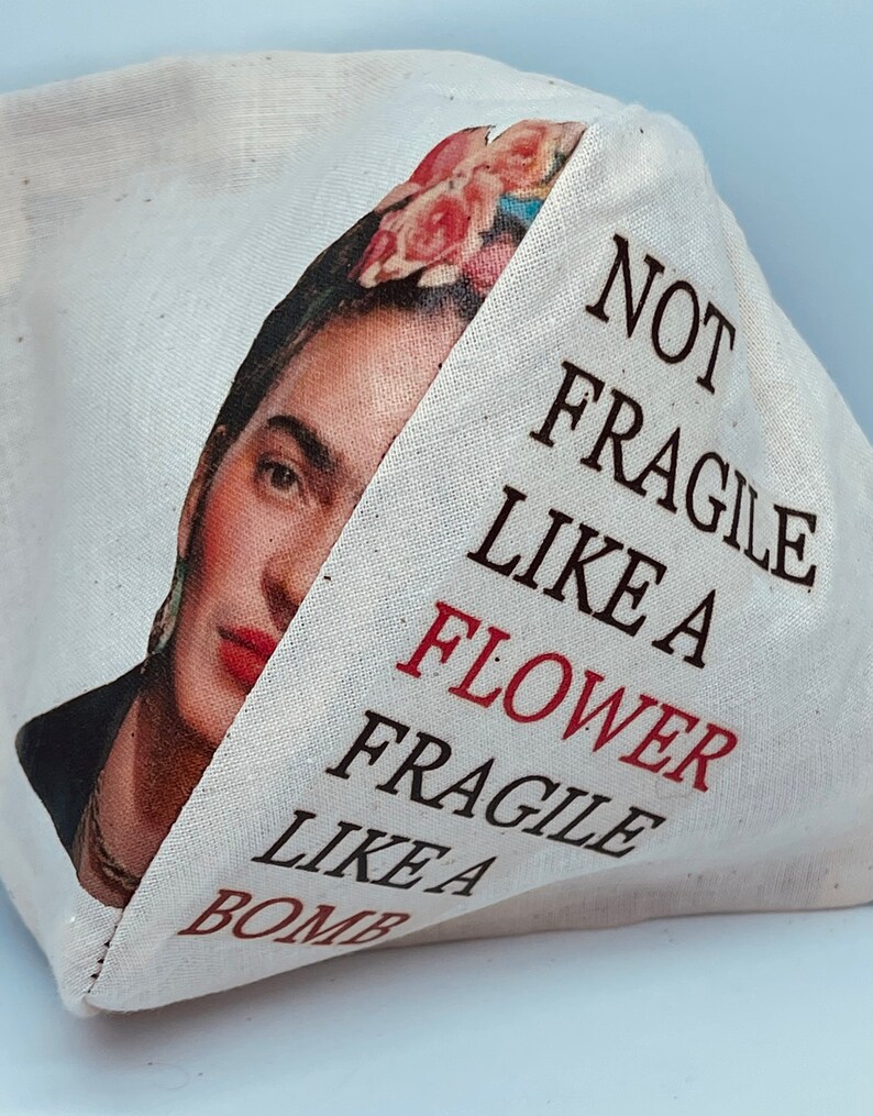 以下が含まれることがあります： フリダ・カーロのカラフルな画像と「Not Fragile Like a Flower Fragile Like a Bomb」というテキストが書かれた白いキャンバス地のトートバッグです。