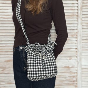 K&ouml;nnte beinhalten: Eine schwarz-wei&szlig; karierte Kordelzugtasche mit passendem Riemen. Die Tasche ist ein kleiner, l&auml;ssiger Stil mit gerafftem Oberteil und einem kleinen Anh&auml;nger. Die Tasche wird &uuml;ber der Schulter getragen.