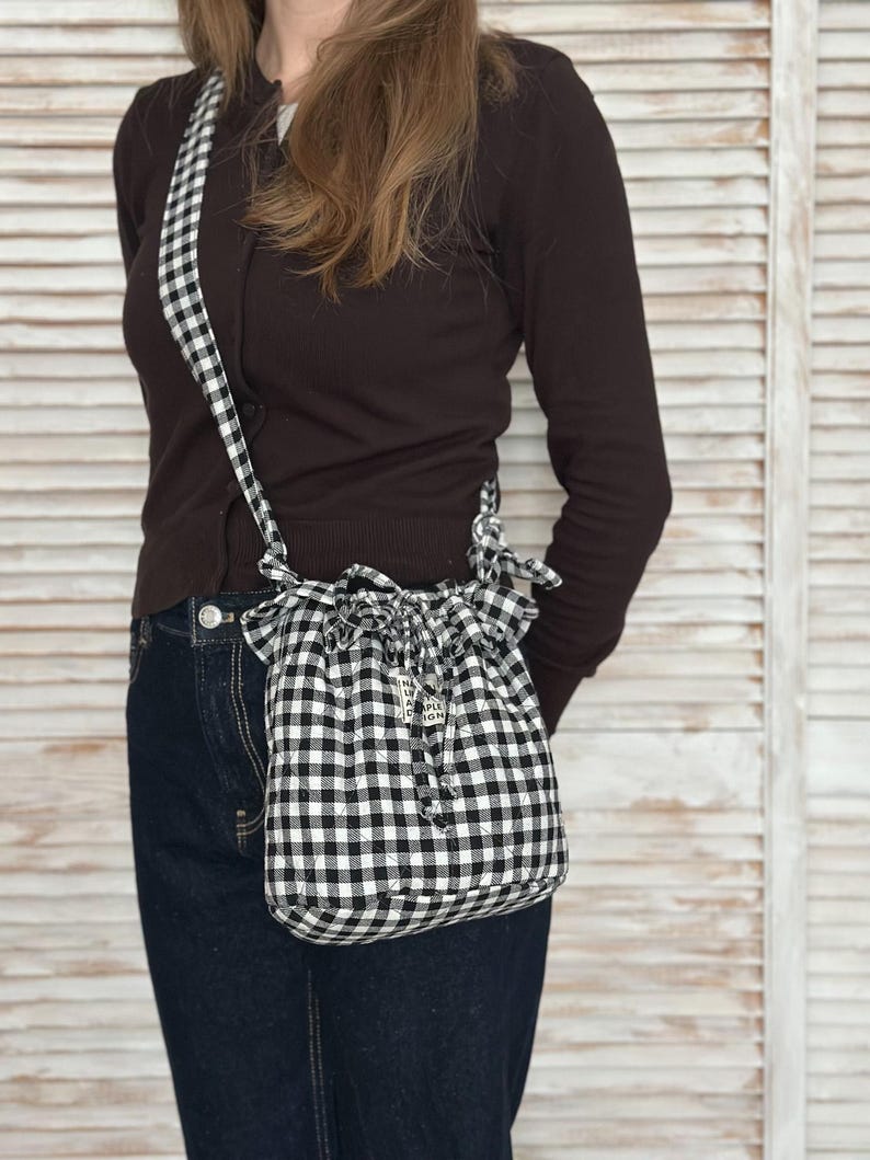 K&ouml;nnte beinhalten: Eine schwarz-wei&szlig; karierte Kordelzugtasche mit passendem Riemen. Die Tasche ist ein kleiner, l&auml;ssiger Stil mit gerafftem Oberteil und einem kleinen Anh&auml;nger. Die Tasche wird &uuml;ber der Schulter getragen.