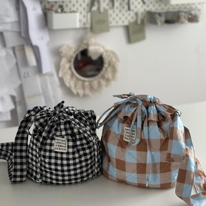 K&ouml;nnte beinhalten: Zwei Kordelzugbeutel auf einer wei&szlig;en Oberfl&auml;che. Einer ist schwarz-wei&szlig;-kariert, der andere braun, blau und beige kariert. Beide Beutel haben ein Etikett mit der Aufschrift "NATURAL LIFESTYLE A SIMPLE DESIGN".