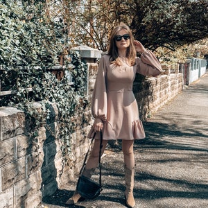 Puede incluir: Una mujer con un vestido marr&oacute;n claro de manga larga y dobladillo con volantes. Lleva botas de vaquero marrones y gafas de sol. Lleva un bolso de mano de cuero negro.