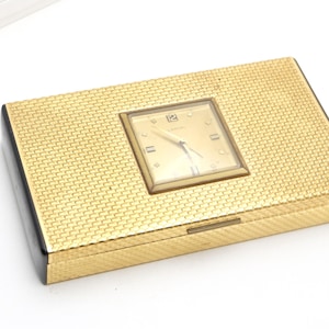 May include: A rectangular gold-tone metal box with a textured basketweave pattern on the lid. A square analog clock is embedded in the center of the lid, featuring a gold face, gold hands, and the text LANCEL printed below the number 12.