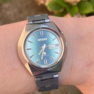 Op de afbeelding: Een zilverkleurig Orient-polshorloge met een ronde, blauwgroene wijzerplaat met goudkleurige uurmarkeringen en wijzers. Het horloge heeft een dag-datumvenster op de drie-uurpositie, met de tekst FRI 18, en een metalen schakelarmband.