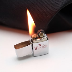 Könnte beinhalten: Ein silberner Metallfeuerzeug mit dem Wort "Winston" in Rot aufgedruckt. Das Feuerzeug ist geöffnet, mit einer hell brennenden Flamme. Der Hintergrund ist ein dunkler Stoff.
