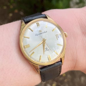 May include: A gold-tone wristwatch with a round, off-white dial featuring gold hour markers, gold hands, and a date window at the three o'clock position. The dial displays the text 'Dynamic Auto' and '33 Jewels'. It has a black leather strap.