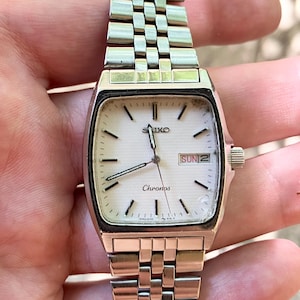 May include: A silver-tone Seiko Chronos wristwatch with a rectangular case and a textured white dial. The watch features black hour markers, silver hands, and a day-date window at the three o'clock position. It has a multi-link metal bracelet.