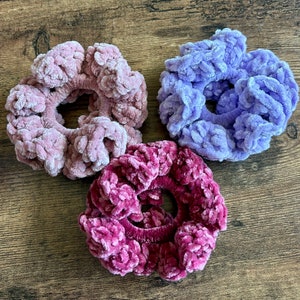 May include: Three crocheted hair scrunchies in different shades of purple. The scrunchies are made of soft, fuzzy yarn and have a gathered, ruffled texture.