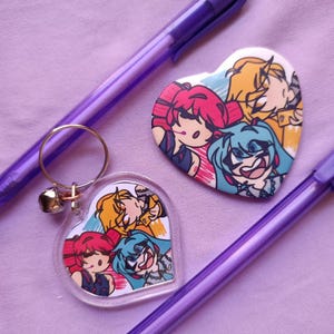 May include: A heart-shaped keychain and button with anime-style character art, featuring red, yellow, and blue hair. Two purple pens are also present on a lavender surface.