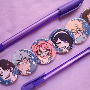 Puede incluir: Seis insignias redondas de personajes con ilustraciones de dibujos animados, dispuestas con dos bolígrafos morados sobre una superficie lavanda. Las insignias presentan varios personajes de estilo anime con diferentes colores de pelo y expresiones. Los bolígrafos son de color morado a juego.