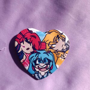 May include: Heart-shaped button featuring three anime-style characters with vibrant hair colors: red, yellow, and blue. The button has a white border and is set against a purple fabric background.