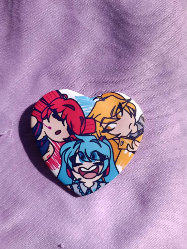 May include: Heart-shaped button featuring three anime-style characters with vibrant hair colors: red, yellow, and blue. The button has a white border and is set against a purple fabric background.