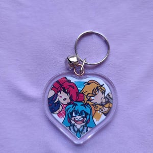May include: A heart-shaped keychain featuring a colorful anime-style illustration of three characters. The keychain has a silver-colored ring and a small bell attached. The background is a light purple fabric.