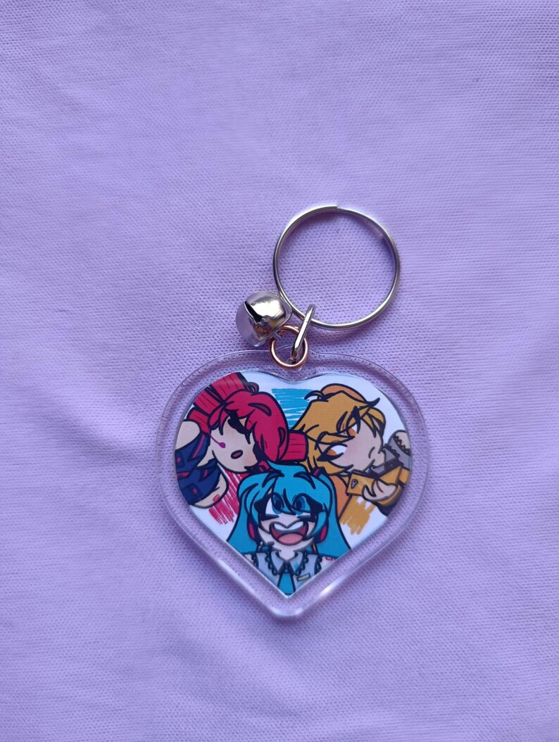 May include: A heart-shaped keychain featuring a colorful anime-style illustration of three characters. The keychain has a silver-colored ring and a small bell attached. The background is a light purple fabric.