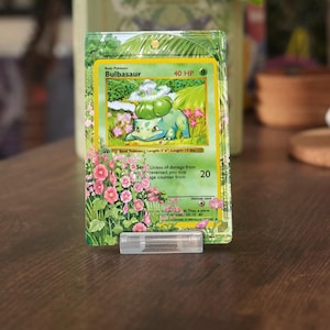 Puede incluir: Un expositor de tarjetas coleccionables de acrílico transparente con un borde decorativo de flores rosas y follaje verde. El marco rectangular tiene un orificio en la parte superior para colgarlo y se apoya en un soporte de plástico. Texto CARD NOT INCLUDED.