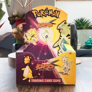 Könnte beinhalten: Eine versiegelte Pokémon Neo Destiny Trading Card Game Box. Die Box zeigt verschiedene Pokémon-Charaktere in Gelb, Grün und Orange. Die Box hat das Pokémon-Logo und den Text "Neo Destiny".