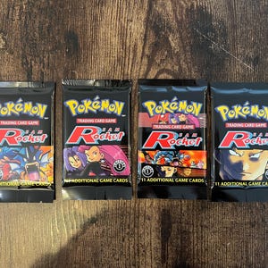 Może przedstawiać: Cztery zapieczętowane pakiety kart kolekcjonerskich Pokémon Team Rocket. Każdy czarny pakiet zawiera logo Pokémon, napisy "Trading Card Game" i "Team Rocket" w kolorze czerwono-białym. Każdy pakiet zawiera 11 dodatkowych kart do gry.