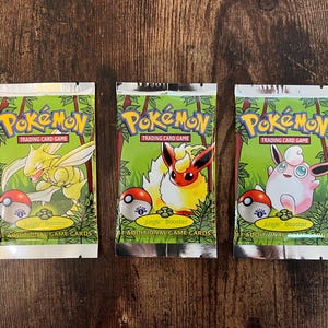 Puede incluir: Tres paquetes sellados de Pokémon Jungle Booster. Cada paquete presenta un personaje Pokémon diferente, incluyendo una criatura amarilla parecida a un insecto, una criatura parecida a un zorro de color naranja y amarillo fuego, y una criatura rosa y redonda.