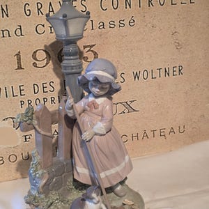 May include: A porcelain figurine featuring a child in a dusty pink dress and blue bonnet standing by a gray street lamp and a wooden fence. A small gray and white cat sits at the base of the lamp, and the child holds a long wooden broom.