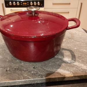 May include: A burgundy cast iron Dutch oven with a lid and side handles. The pot is sitting on a gray marble-like surface. The kitchen setting includes a stove and cabinets.