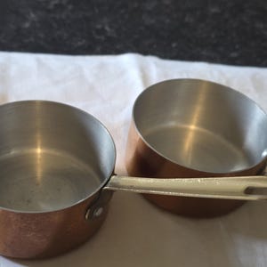 May include: Two copper-coloured saucepans with silver-coloured handles. The pans have a rounded shape and are made of copper with a shiny interior. The handles are attached with rivets. The pans are on a white surface.