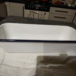 May include: A rectangular white enamel baking dish with a dark blue rim. The dish is empty and sits on a white lace tablecloth. The kitchen background includes a cooker and cabinets.